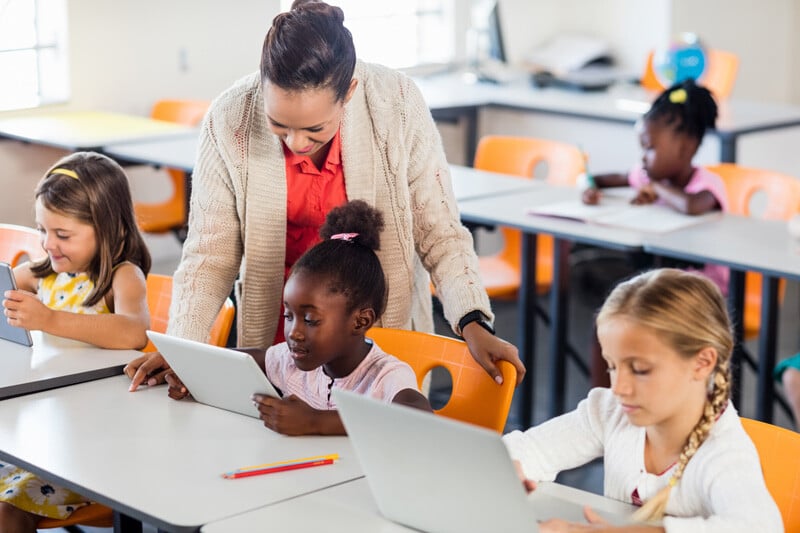 Young children in a classroom working with teacher on various devices.