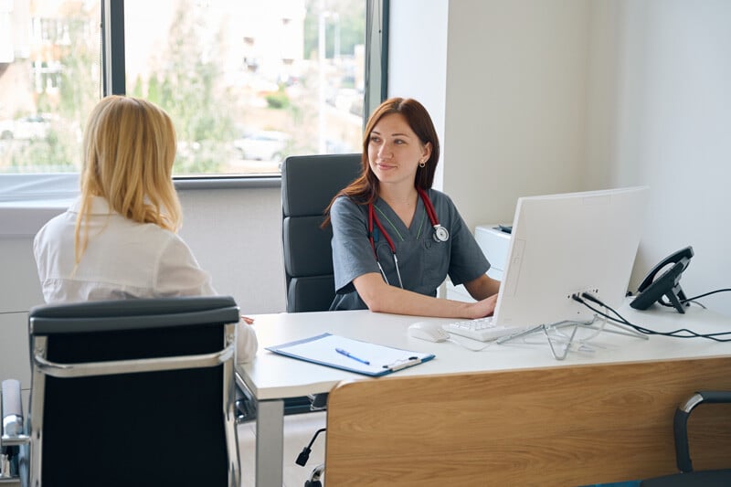 A healthcare professional typing on a computer while speaking with a patient.